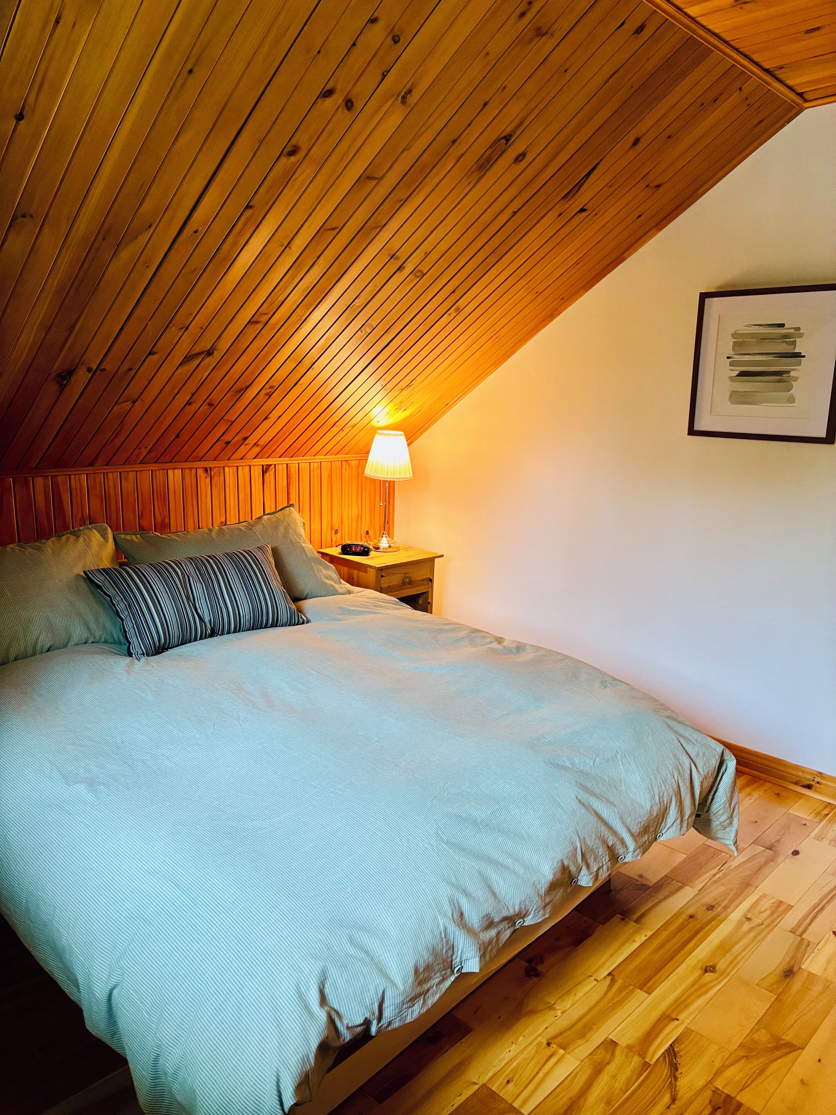 A bedroom with a sloped ceiling, a bed with a striped bedspread, a bedside table with a lamp, and a framed picture on the wall.