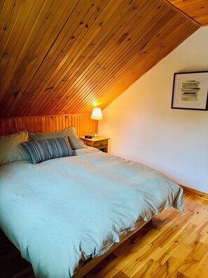 Ein Schlafzimmer mit schrägen Wänden, einem Bett mit gestreifter Bettdecke, einem Nachttisch mit Lampe und einem gerahmten Bild an der Wand.