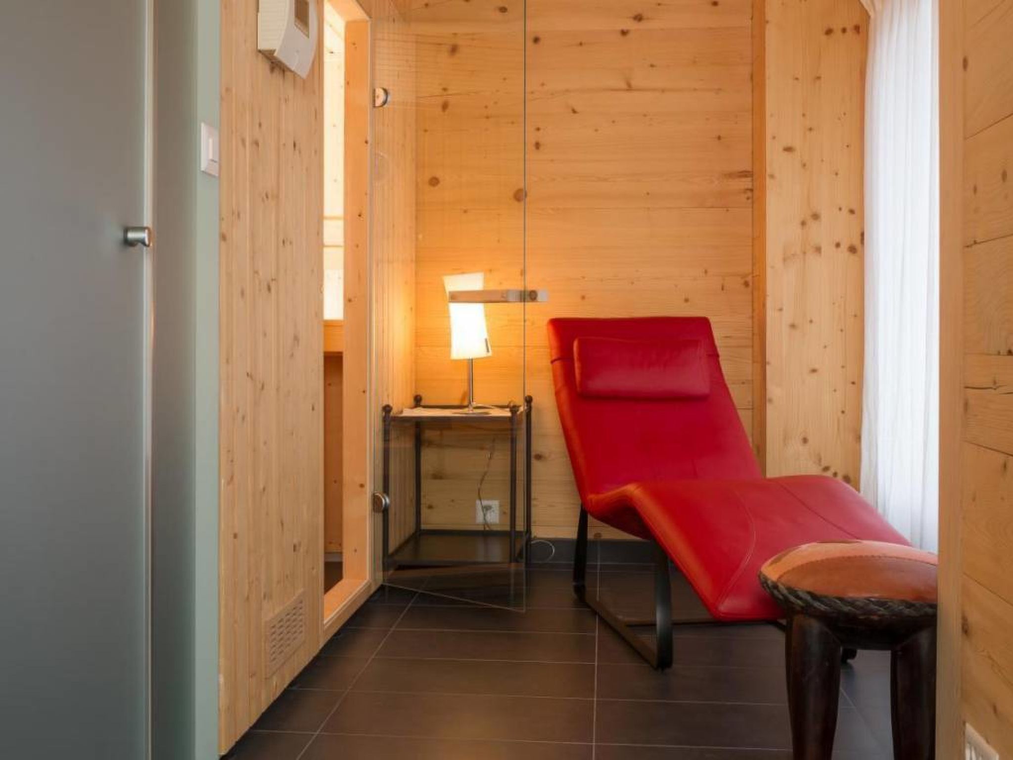 A wooden room with a red massage chair, a small table with a lamp, and a door.