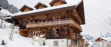 A wooden chalet with a balcony, surrounded by snow-covered ground and mountains.