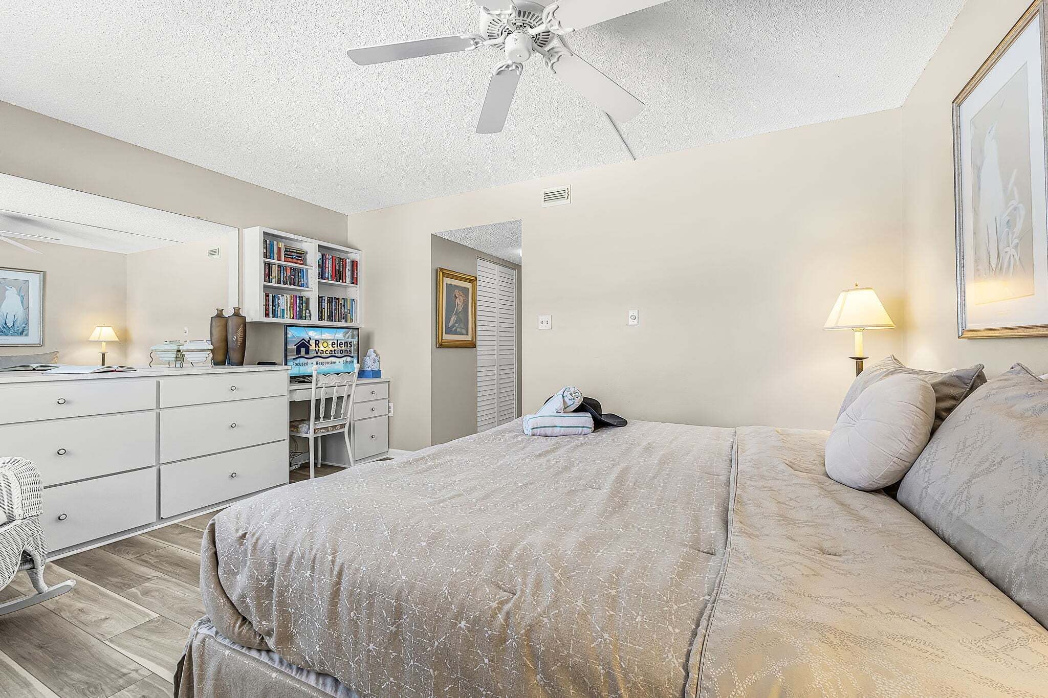 A bedroom with a bed, a desk with a computer, a bookshelf, and a ceiling fan.