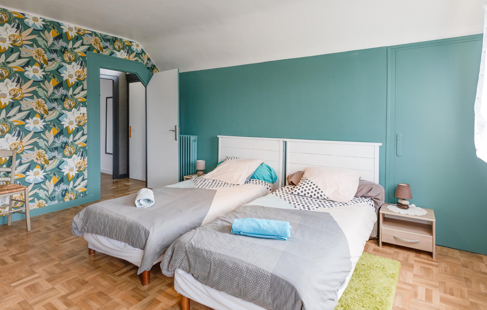 A bedroom with two beds, a wooden floor, a teal accent wall, and a floral wallpaper on one side.