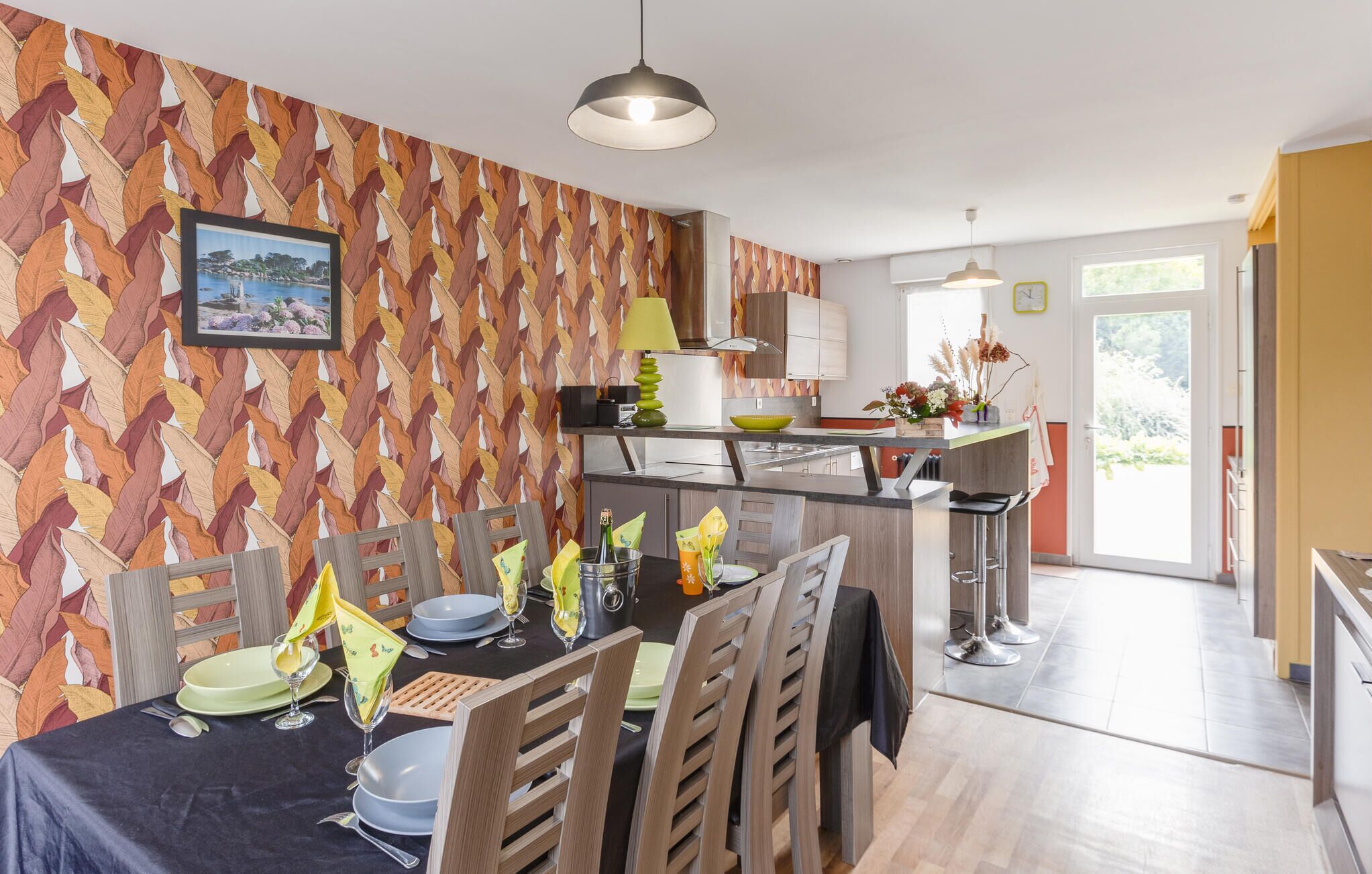 Una cocina moderna con un papel tapiz decorativo, una zona de comedor con sillas de madera y una mesa preparada para dos personas, y vistas al exterior a través de un ventanal.