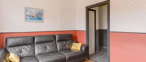 A living room with a sectional sofa, a glass coffee table, a framed picture on the wall, and a door leading to another room.