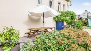 Een terras met een houten tafel en een witte parasol, omgeven door groen en een gebouw.