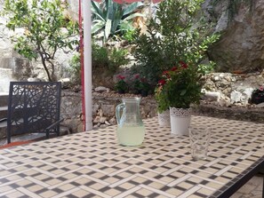 Ein draußen stehender Tisch mit karierter Tischdecke, einer Wasserkaraffe, einem Glas und Topfpflanzen.
