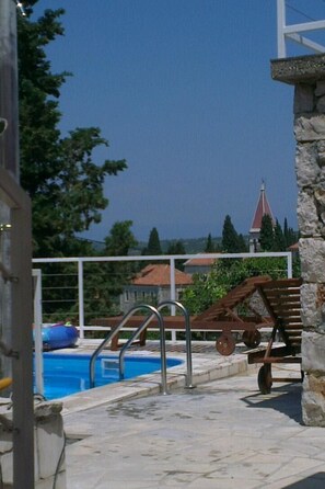 Ein Schwimmbecken mit Metallleitern, einer Steinmauer und Blick auf eine Kirche mit rotem Dach.