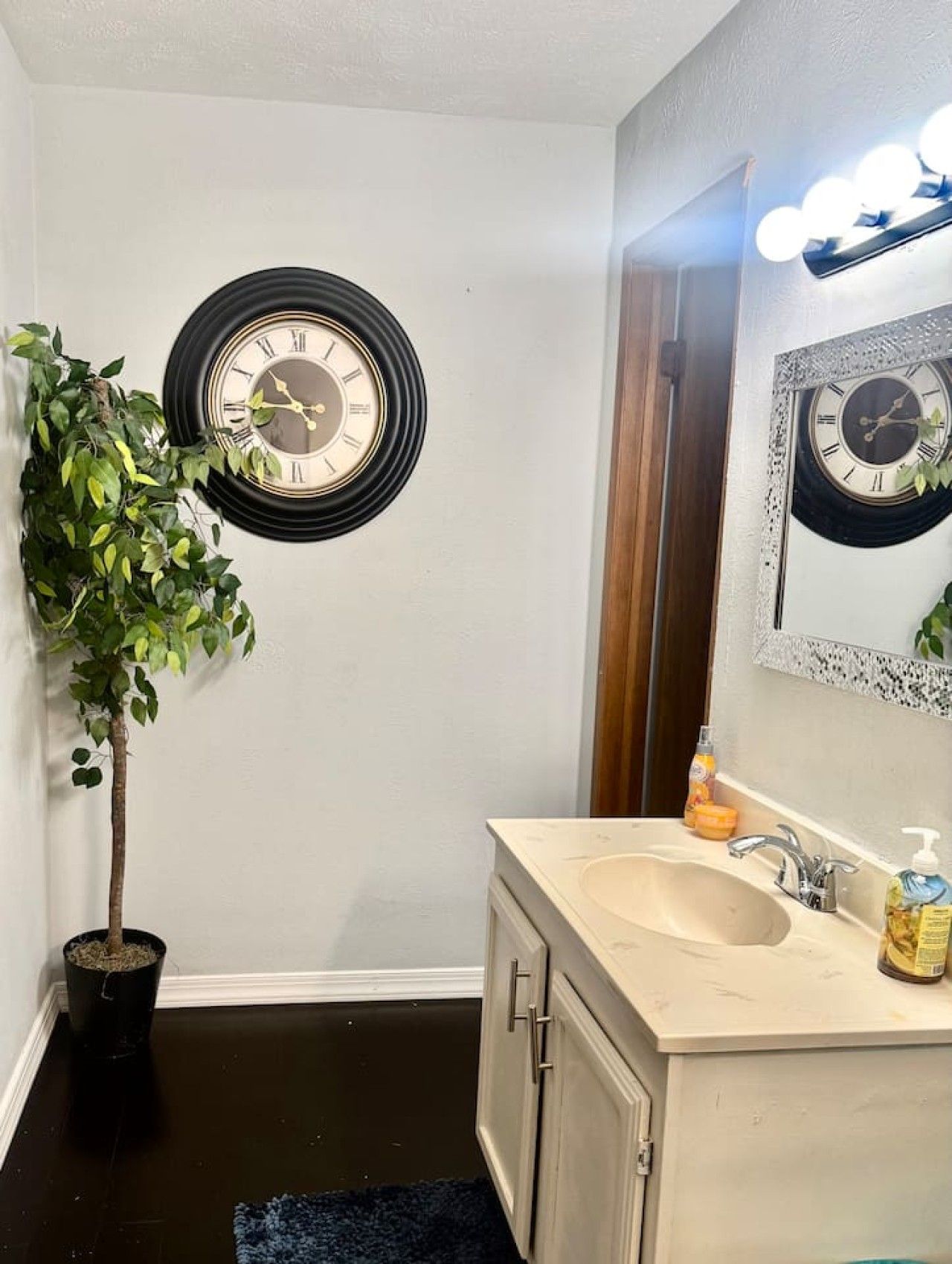 Une salle de bain avec une horloge murale ronde, une plante en pot, un lavabo avec des articles de toilette et un miroir avec éclairage.