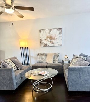 Un salon moderne avec un ventilateur de plafond, une table basse ronde, deux canapés gris, des coussins à motifs, une lampe sur pied et un grand tableau abstrait accroché au mur.