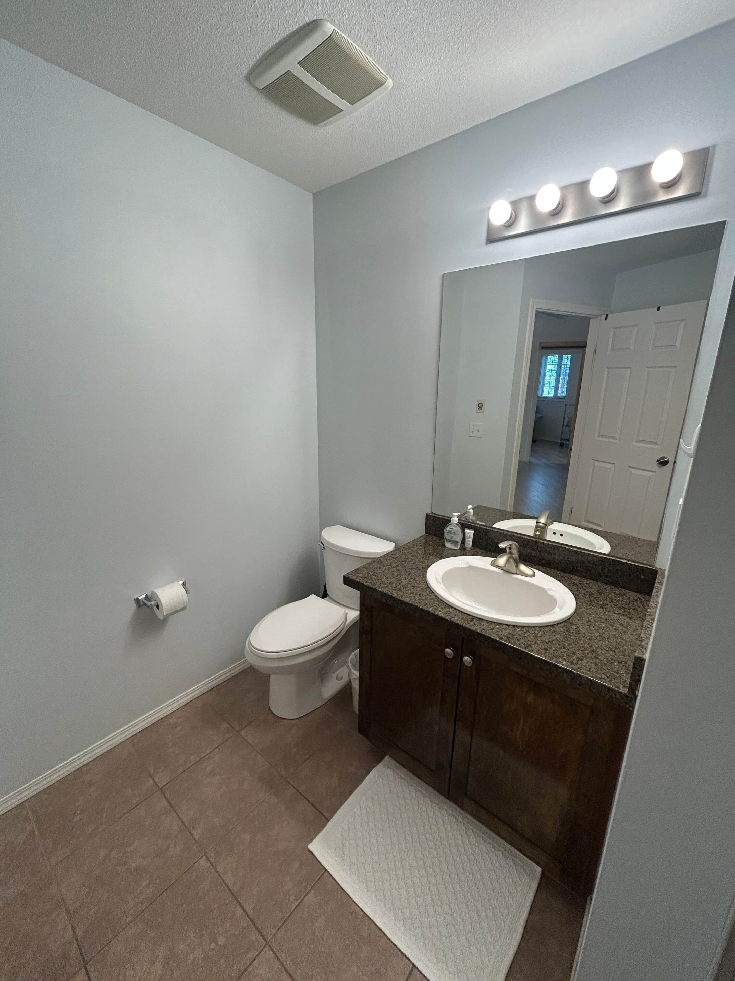 A bathroom with a toilet, sink, vanity, mirror, and a light fixture above the mirror.