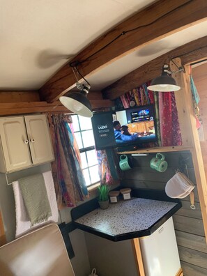 A compact kitchen with wooden beams, a small countertop, a refrigerator, and a television displaying a show.