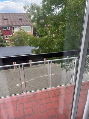 Ein Balkon mit Metallgeländer, Fliesenboden und Blick auf ein Wohngebiet mit Häusern und Bäumen.