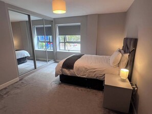Ein Schlafzimmer mit einem Bett, Nachttisch und Lampe. Große Fenster bieten einen Ausblick nach draußen.