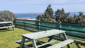 Ein Picknickbereich mit Holzbänken und -tisch, umgeben von einem grünen Zaun, mit Blick auf einen Strand und den Ozean.