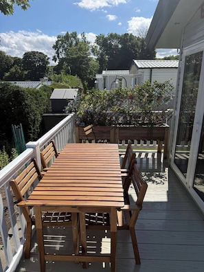 Eine Holzterrasse mit Tisch und Stühlen, umgeben von einem weißen Geländer, und eine Glastür zu einem überdachten Bereich.