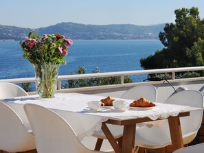 A table set with a croissant, two cups, and a vase of flowers, overlooking a body of water with a mountainous backdrop.