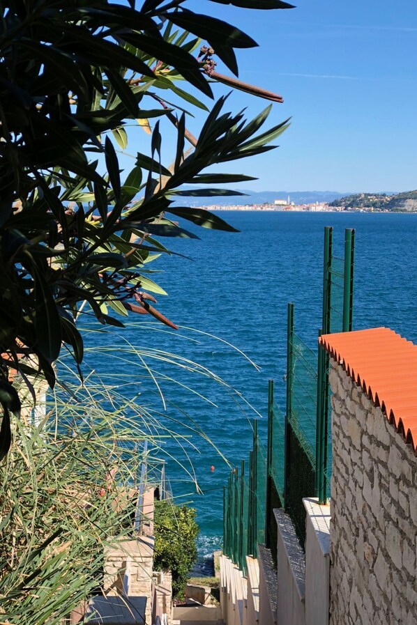 A coastal view with a clear blue sea, a stone building with a red roof, and a distant cityscape.