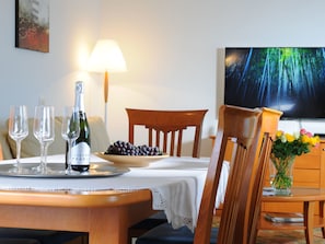 A dining area with a table set for two, a bottle of wine, glasses, a bowl of grapes, and a television displaying a forest scene.
