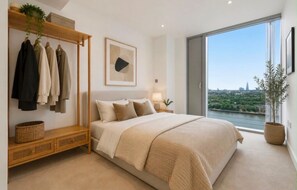 A bedroom with a large bed, a wardrobe, a bedside table, a lamp, and a view of a cityscape through a large window. - Canary Wharf Skyline Retreat (London)