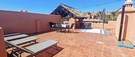 En tagterrasse med et bord og stole, et overdækket siddeområde og udsigt til de omkringliggende bygninger.
