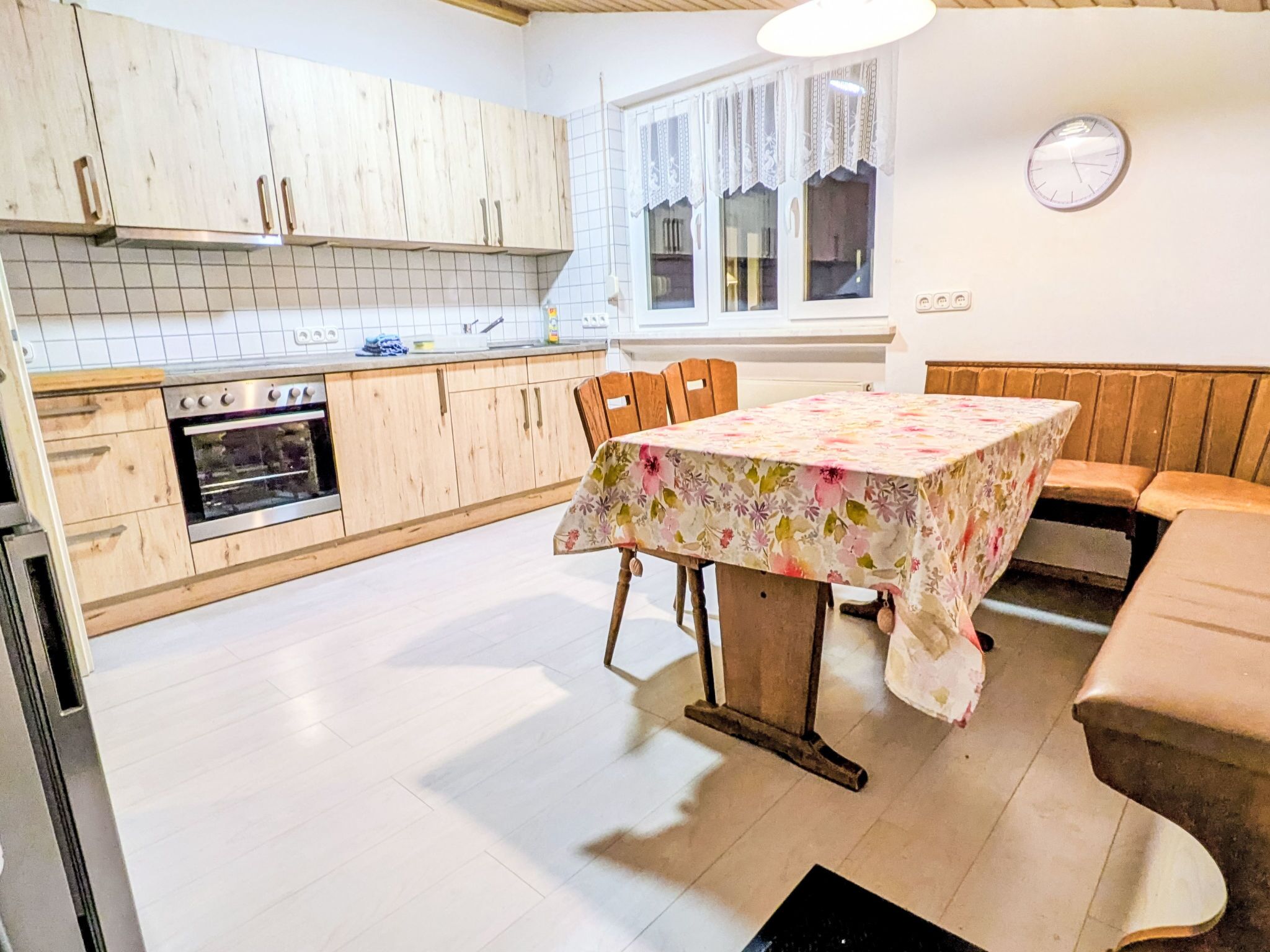 Eine Küche mit Holzschränken, einem Esstisch mit Blumenmuster-Tischdecke und einer Wanduhr.