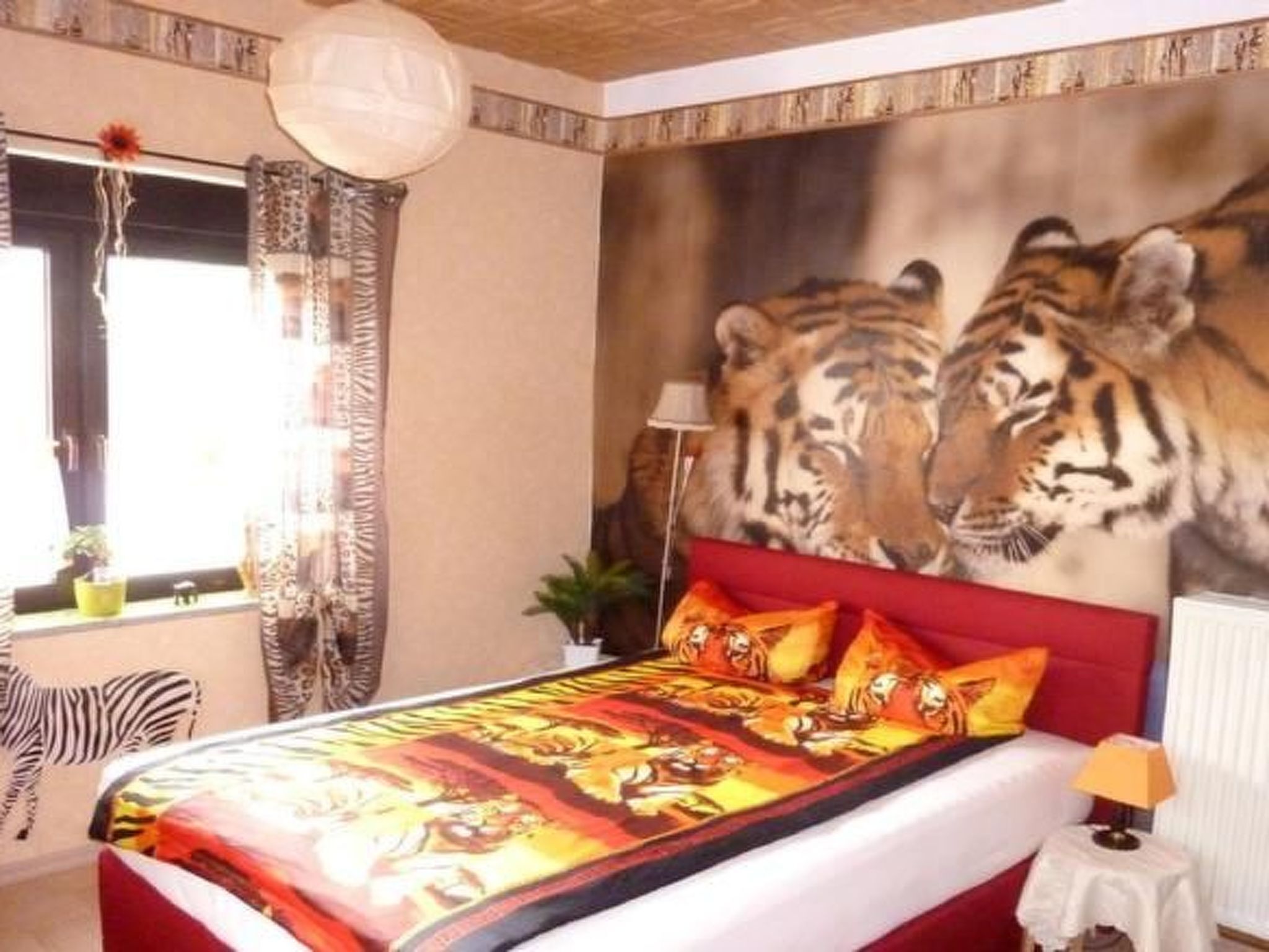 A bedroom with a large tiger mural on the wall, a zebra-patterned chair, and a bed with a red headboard.