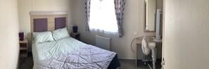 Une chambre à coucher comprenant un lit, une table de chevet, un radiateur, un ventilateur sur pied et une fenêtre avec des rideaux.