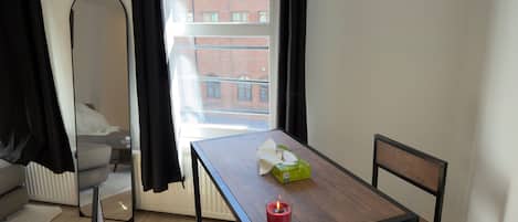 Une pièce avec un lit, une table sur laquelle est posée une bougie, une boîte et une vue sur un immeuble par la fenêtre.