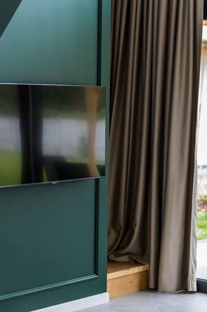 A room with a large flat-screen TV mounted on a green wall, a wooden bench, and a brown curtain partially drawn to the side. - in Niechorze mit Großem Grill by Interhome (Niechorze)