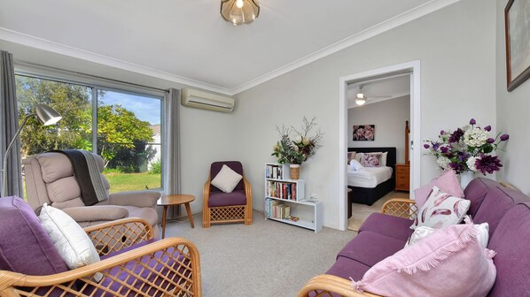 A living room with a sofa, armchair, and a small table. There is a bookshelf with books and a vase with flowers. A bed is visible in the background. - Morpeth Cottage | In the hear of Morpeth (Morpeth)