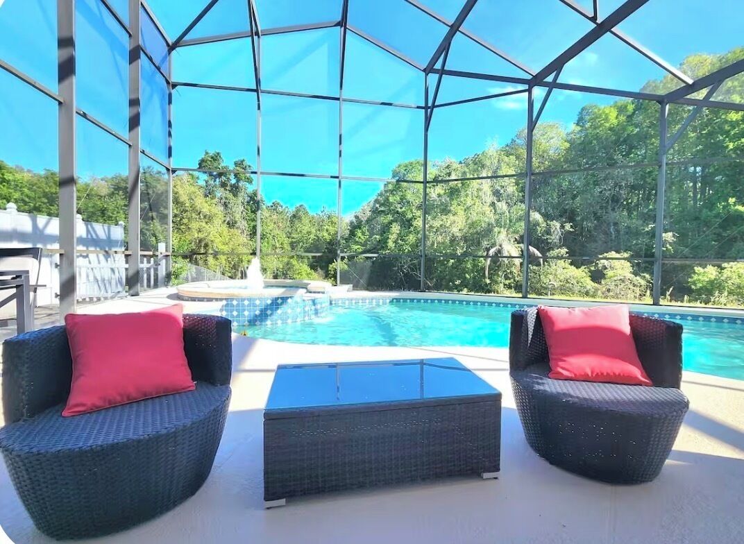 A covered outdoor area with a pool, two wicker chairs with red cushions, and a table.