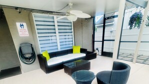 Un área de estar al aire libre con techo, que incluye un sofá y dos sillones redondos, un aire acondicionado instalado en la pared y un letrero que indica supervisión de adultos para niños.