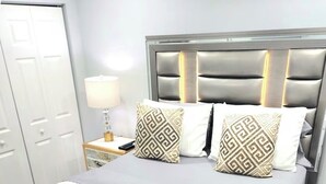 Una cama bien tendida con una colcha gris, dos almohadas estampadas y una manta blanca.