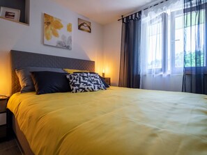 Ein Bett mit gelber Bettdecke und mehreren Kissen, ein Nachttisch mit Lampe sowie ein Fenster mit Vorhängen.
