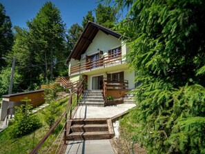 Eine zweistöckige Chalet mit einer Holztreppe, die zu einem Balkon führt, umgeben von üppiger Vegetation.