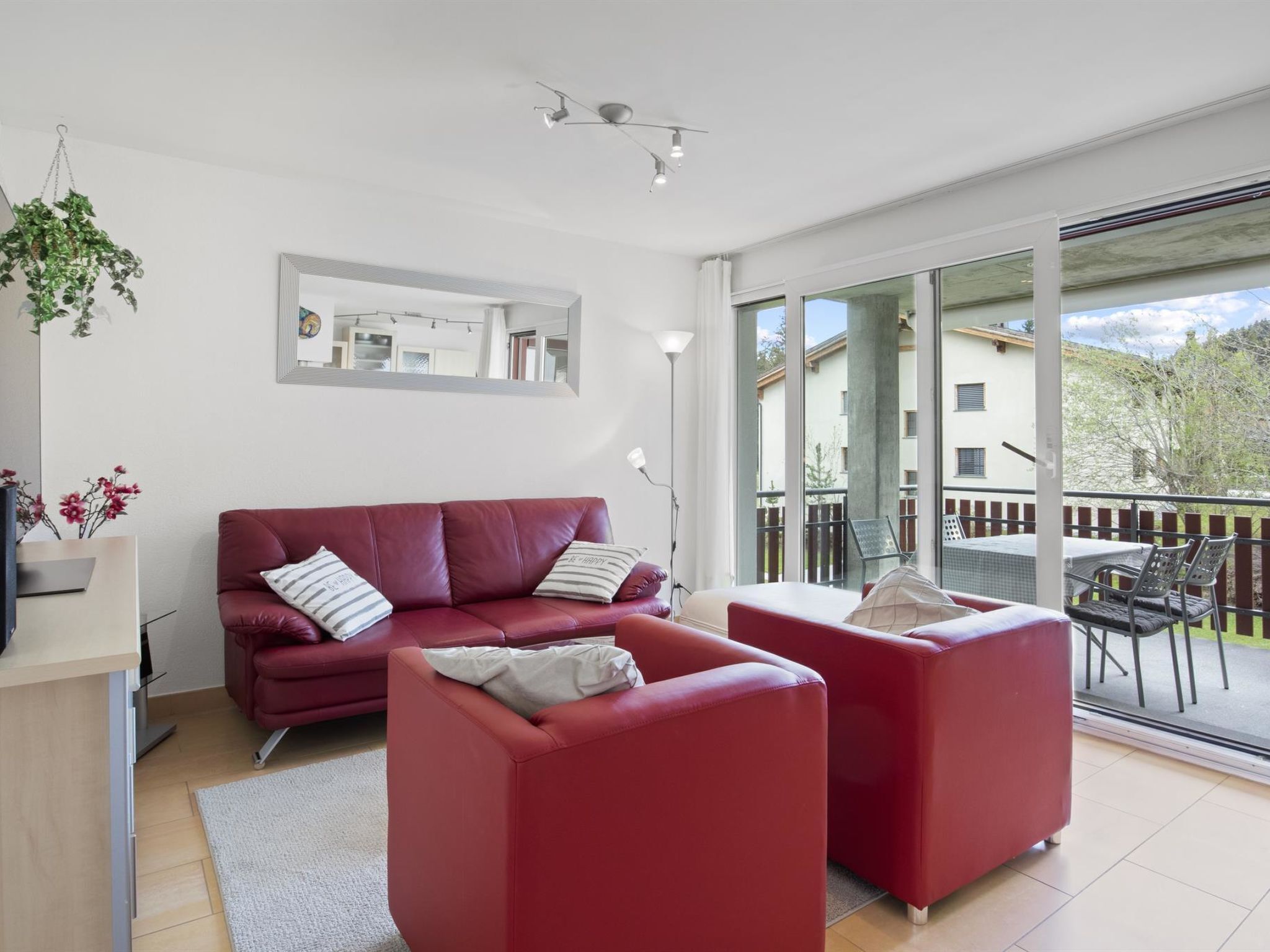 En moderne stue med en rød sofa, en mindre rød lænestol og en glasdør til et udendørs område.