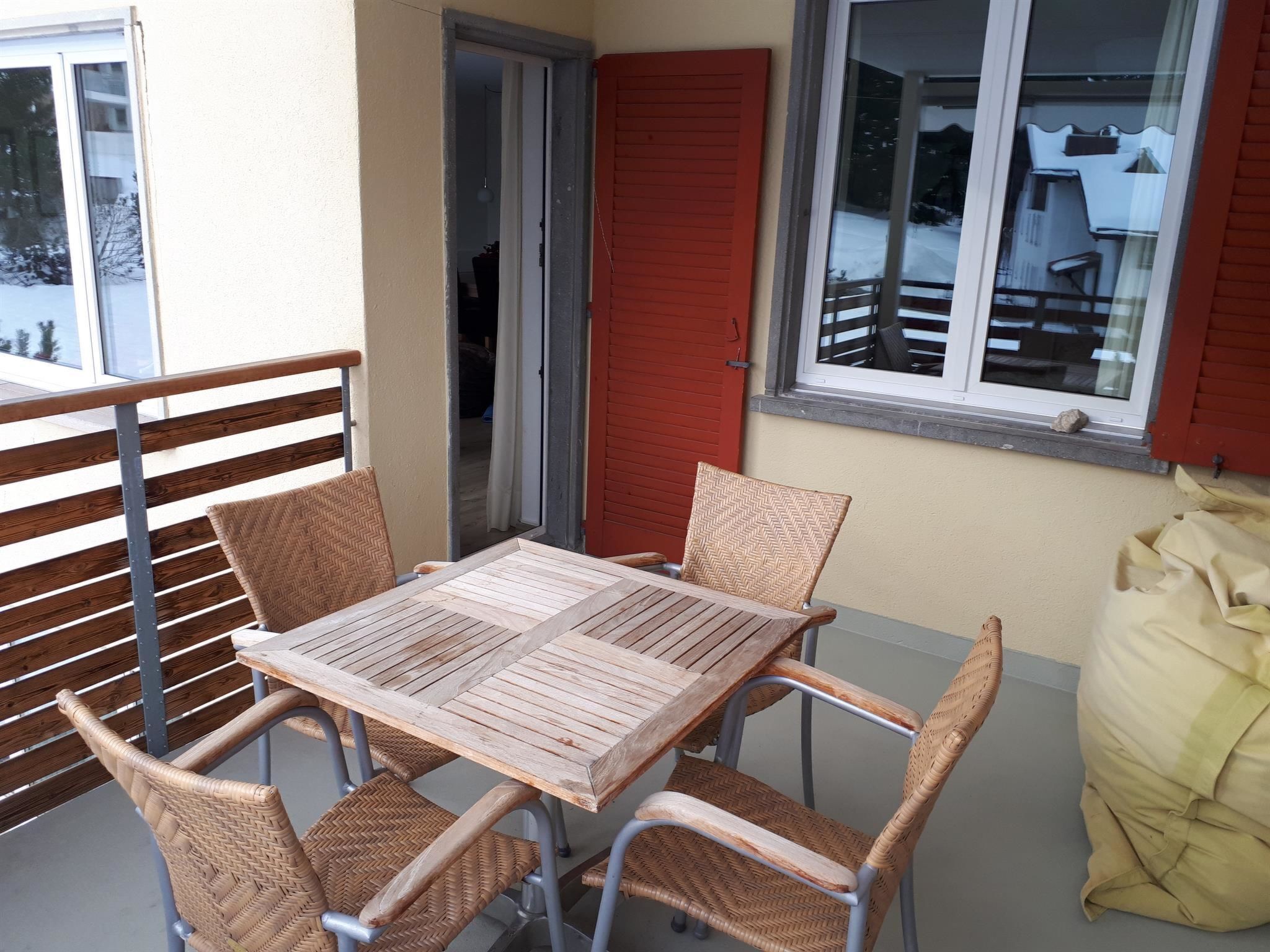 A balcony with a table and four wicker chairs, a red shutter door, and a window overlooking a snowy landscape.