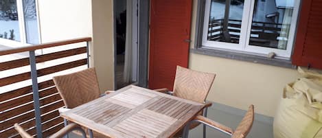 A balcony with a table and four wicker chairs, a red shutter door, and a window overlooking a snowy landscape.