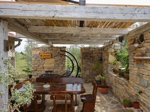 An outdoor patio with a stone wall, a wooden table, and chairs. There is a sign that reads 'Bratovici'. - Olive Garden Paljka by Interhome (Donje Rastane)