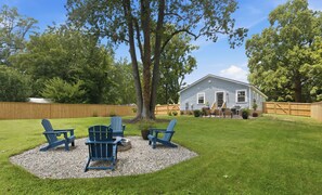 Een achtertuin met een houten terras, drie Adirondack-stoelen, een kleine vuurkuil en een huis op de achtergrond.