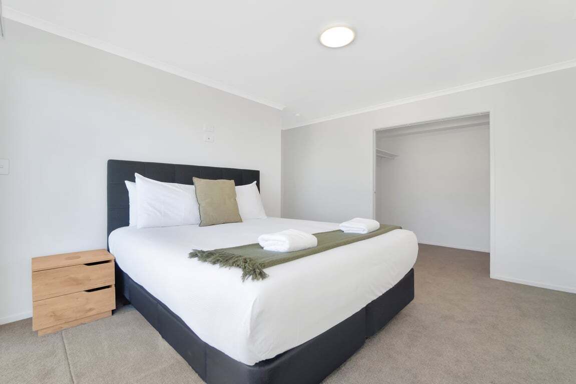 A neatly made bed with white linens and a dark headboard, a small wooden nightstand, and a closet door in the background.