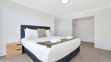 A neatly made bed with white linens and a dark headboard, a small wooden nightstand, and a closet door in the background.