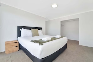 A neatly made bed with white linens and a dark headboard, a small wooden nightstand, and a closet door in the background. - The Seaway Vista (Tauranga)