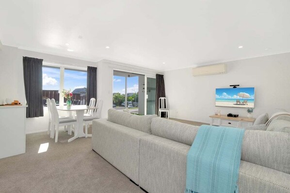 A modern living room with a sofa, a dining table with chairs, and a television on the wall.