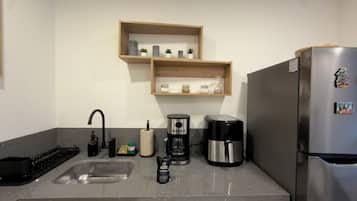 A kitchen with a stainless steel refrigerator, a coffee maker, a sink, and wooden shelves above holding jars and small plants.