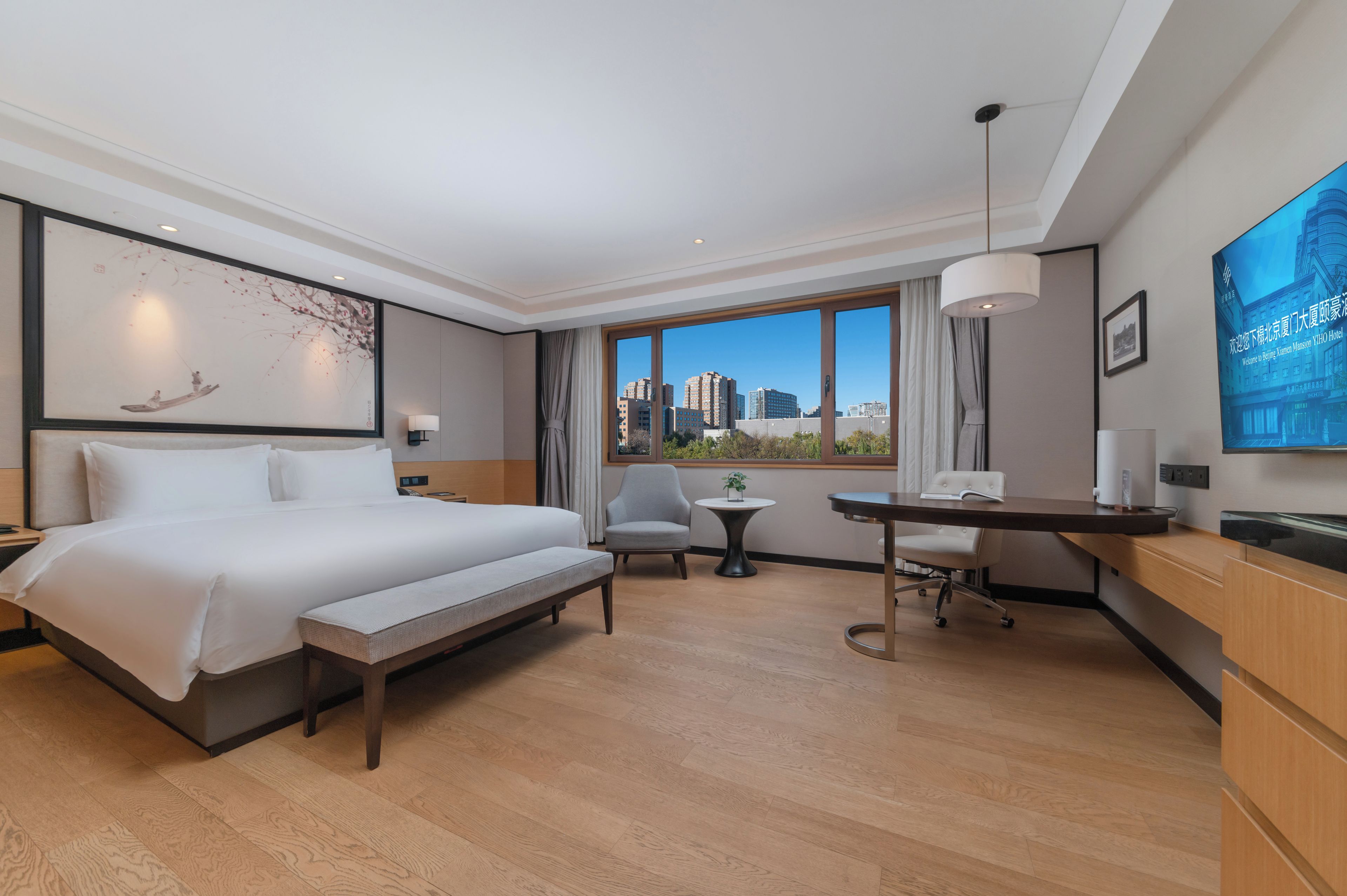 Habitación de hotel con una cama grande, un escritorio, una silla, una mesita, una lámpara y un ventanal con vistas a la ciudad.