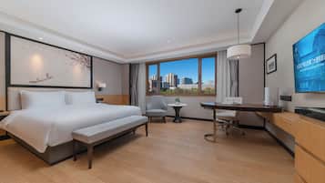 Quarto de hotel com uma cama grande, uma escrivaninha, uma cadeira, uma mesinha, um abajur e uma janela ampla com vista para a cidade.