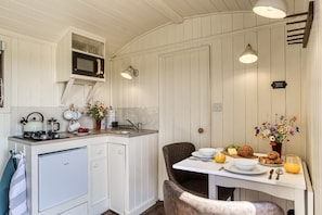 A compact kitchen with a microwave, kettle, and sink, a dining table with a chair, and a small refrigerator.