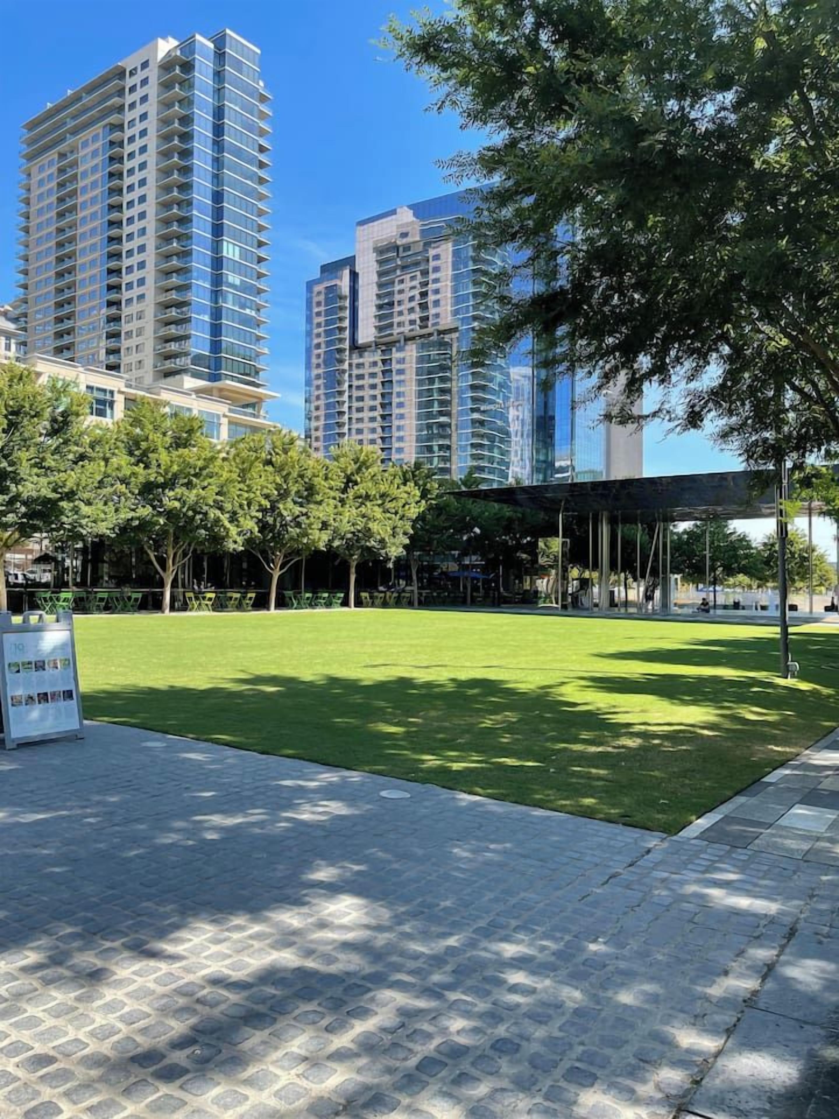 A park with a paved walkway, a covered seating area, and tall buildings in the background.