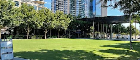 A park with a paved walkway, a covered seating area, and tall buildings in the background.